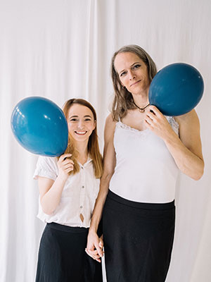Two women in white tops and black skirts holding hands, each holding a blue balloon in their free hand