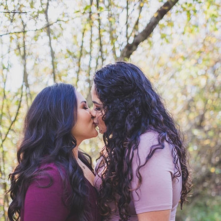 Two Latinx women in pink kissing, backed by soft-lit trees